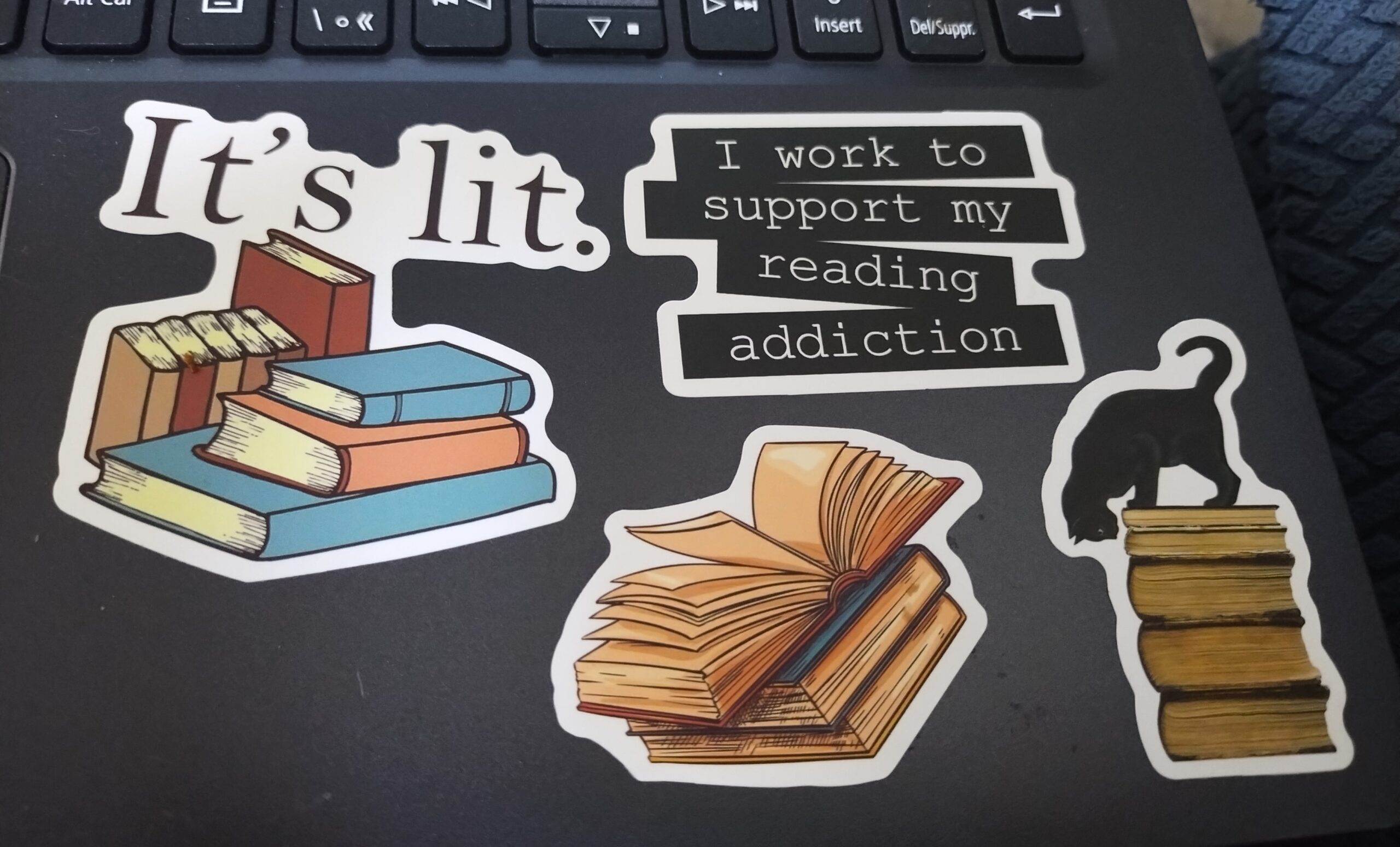 Picture of several bookish stickers on a laptop. One says "I work to support my reading addiction." One says "It's Lit.". One is a black cat standing on top of a pile of books.